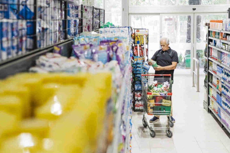 Senior man with shopping cart looking at grocery products in supermarket aisle. Mature male customer looking at groceries in store.