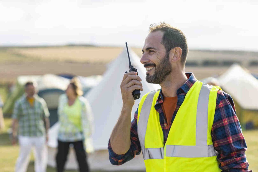 A volunteer wearing a reflective vest and working at a festival in Lindisfarne, North East England. He is talking to someone using a walkie talkie while smiling.