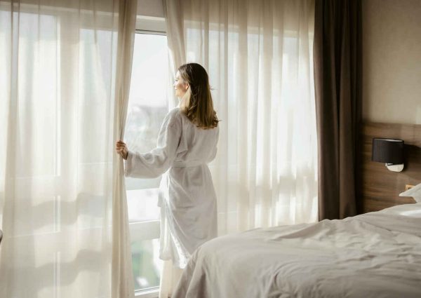 Woman enjoys magnificent view from window in modern apartment or hotel. Young female wearing white bathrobe opening curtains in the morning